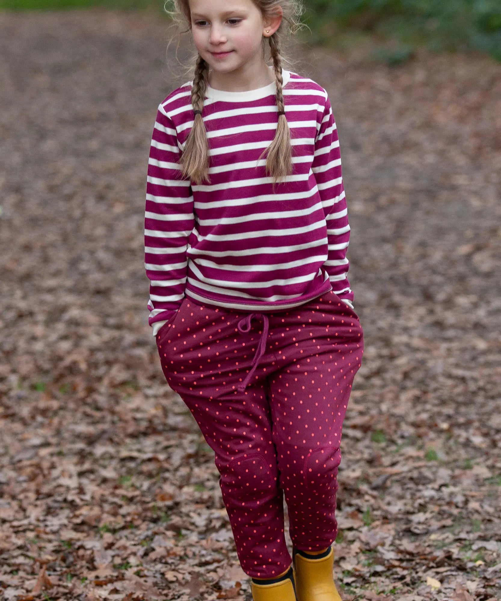 Child wearing the Little Green Radicals autumn polkadot cosy cotton knee patch joggers with a striped berry long sleeve top and yellow gold wellington boots.