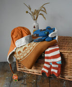 Little Green Radicals gold yellow mountain fair isle snuggly woollen jumper placed on wicker basket with a corduroy flapper hat, bear sherpa gloves and striped socks. 