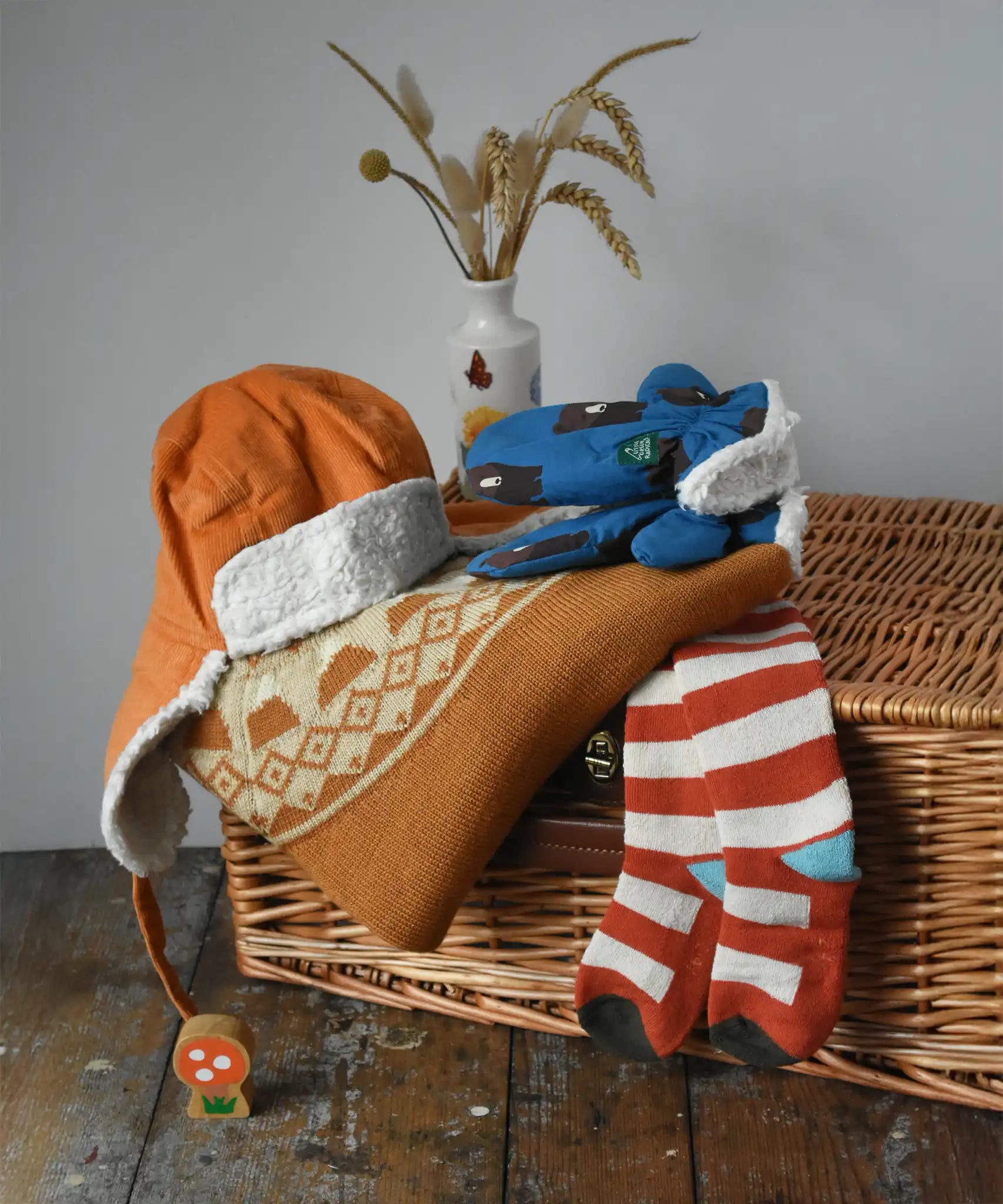 Little Green Radicals gold yellow mountain fair isle snuggly woollen jumper placed on wicker basket with a corduroy flapper hat, bear sherpa gloves and striped socks. 