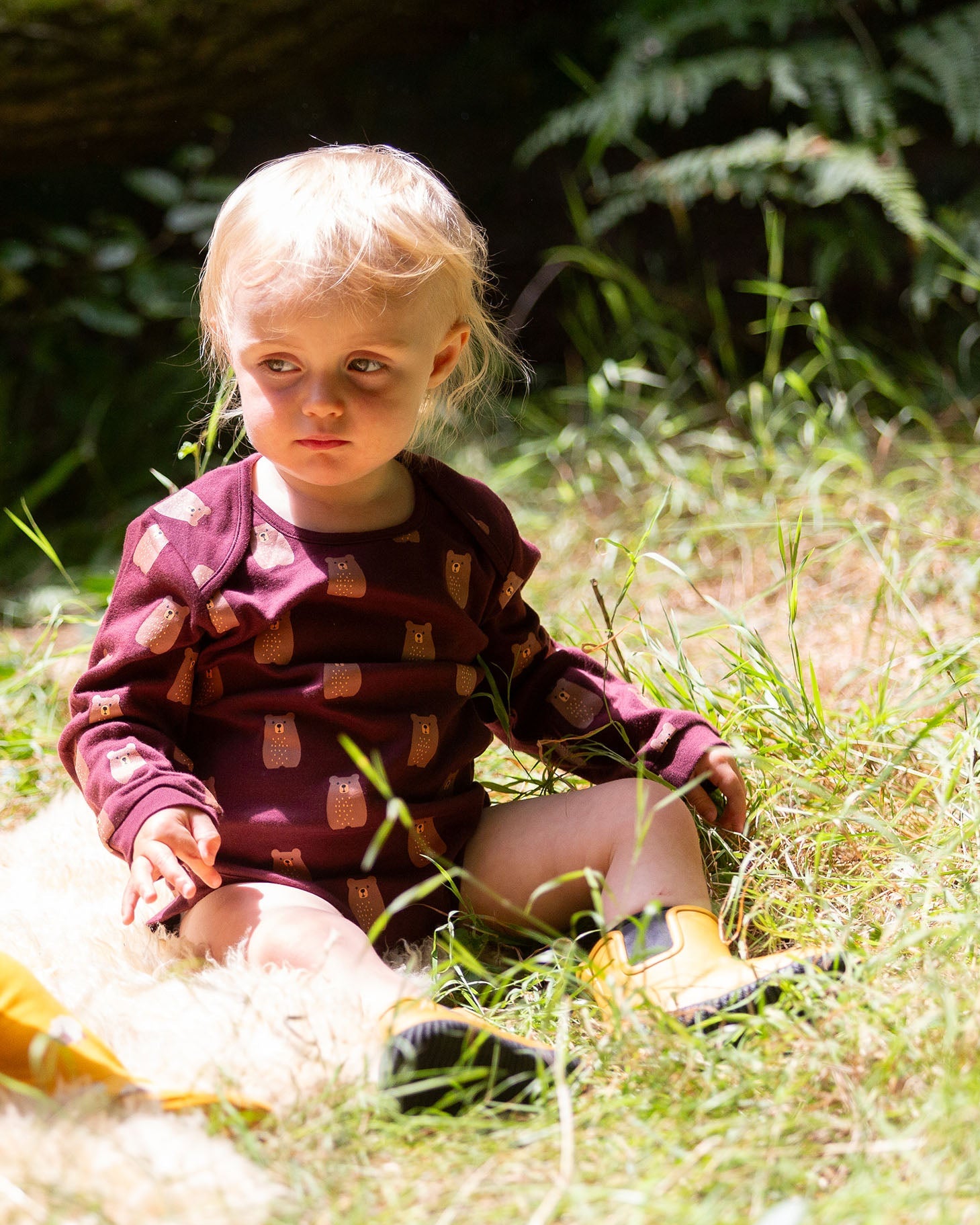 A young child sitting on grass wearing a Little Green Radicals Baby Bodysuit 2 Pack featuring a Brown Bears print. Little Green Radicals offer a range of organic cotton children's clothing available here at Babipur. 