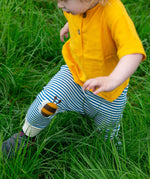 A close up of a child's legs wearing the Little Green Radicals Bees Knees Patch Striped blue and white Joggers. The child is also wearing a yellow gold coloured short sleeved t-shirt whilst running through long green grass.