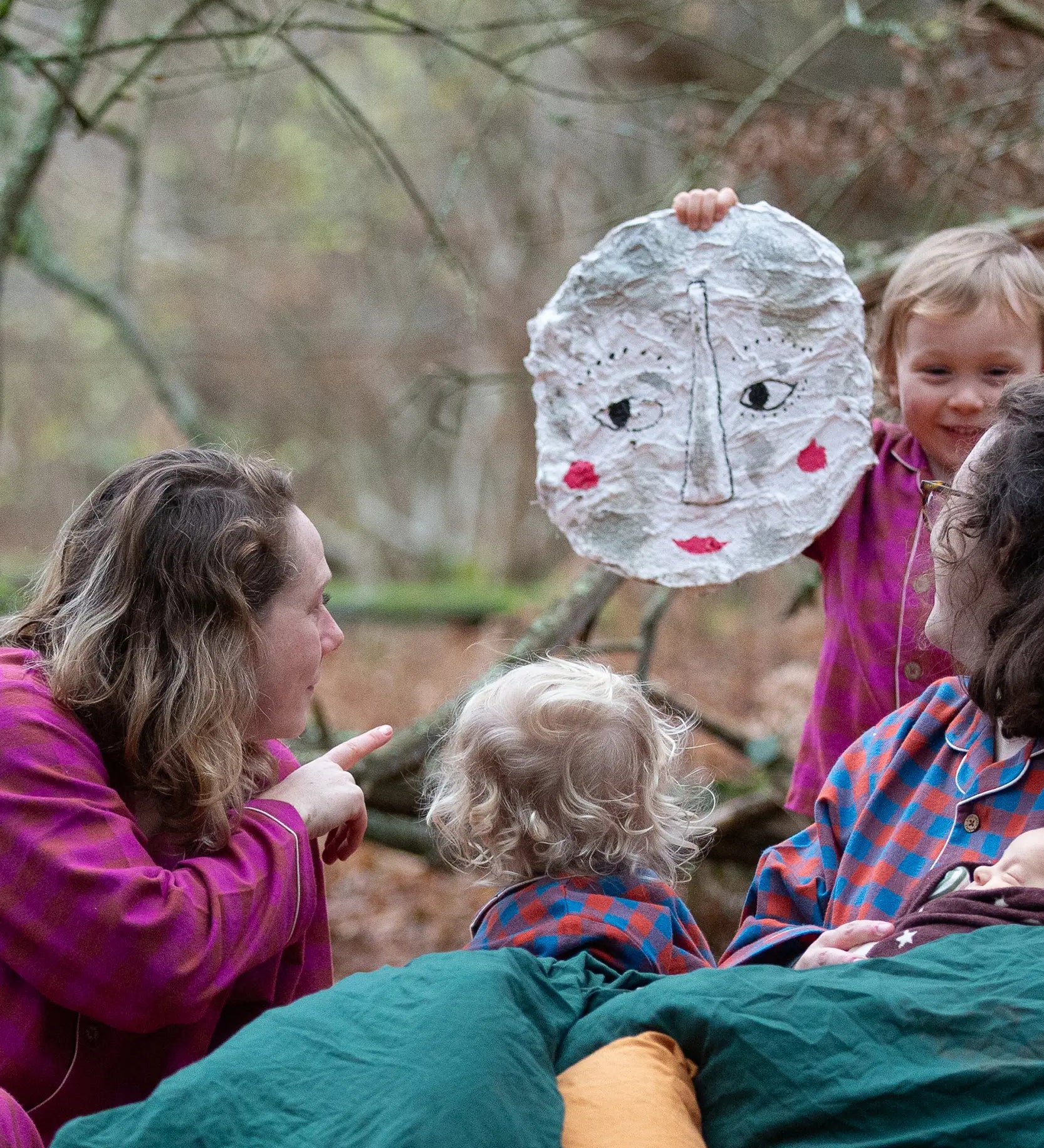 Woman wearing the Little Green Radicals berry check adult classic pyjamas sitting outdoors next to children in Little Green Radicals organic cotton pyjamas.