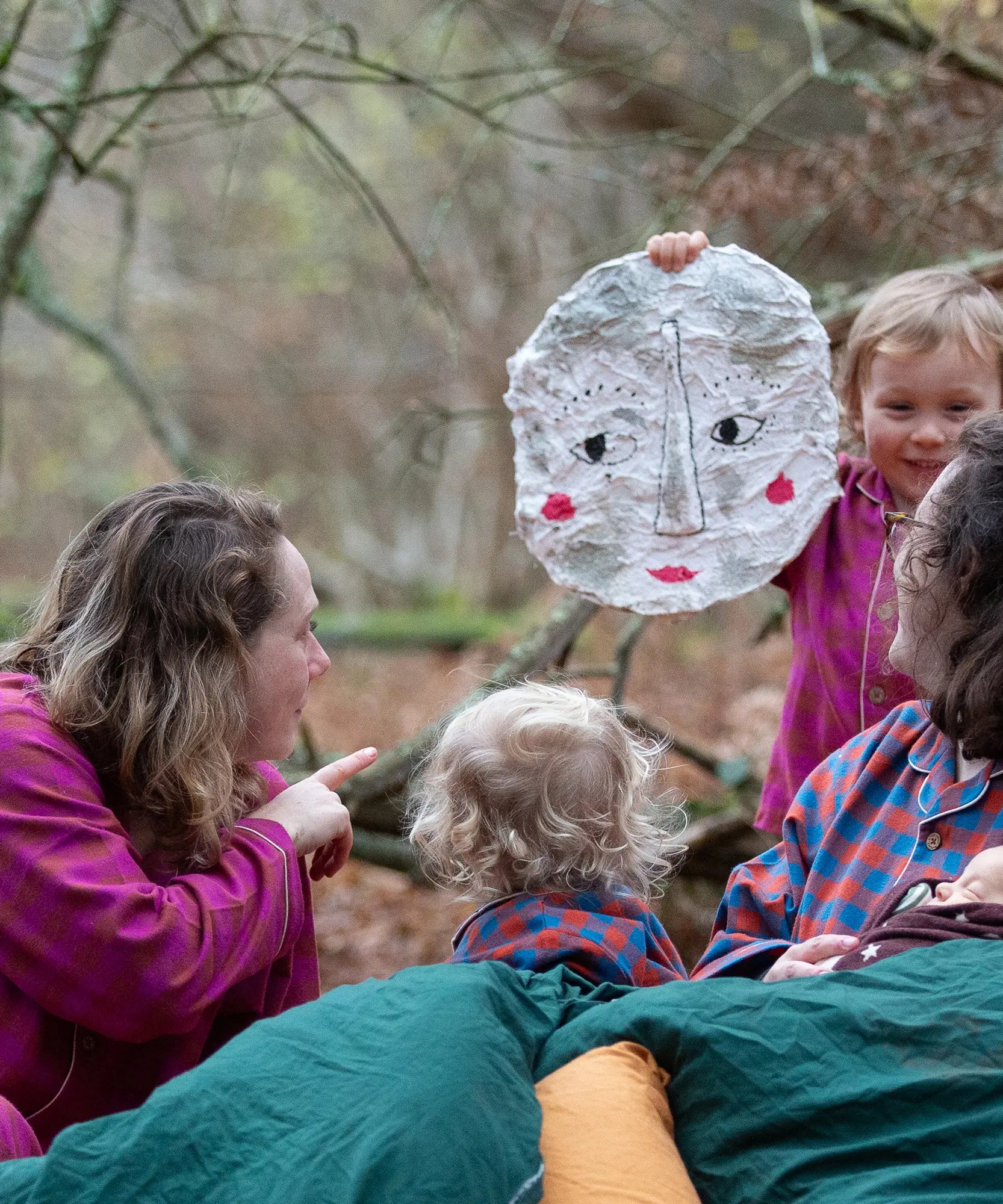 Woman wearing the Little Green Radicals berry check adult classic pyjamas sitting outdoors next to children in Little Green Radicals organic cotton pyjamas.
