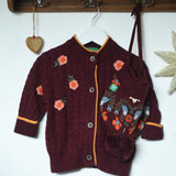 Little Green Radicals organic cotton snuggly knitted cardigan with a berry petal design and yellow edges on a hanger next to a folk birds hat.