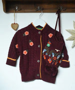 Little Green Radicals organic cotton snuggly knitted cardigan with a berry petal design and yellow edges on a hanger next to a folk birds hat.
