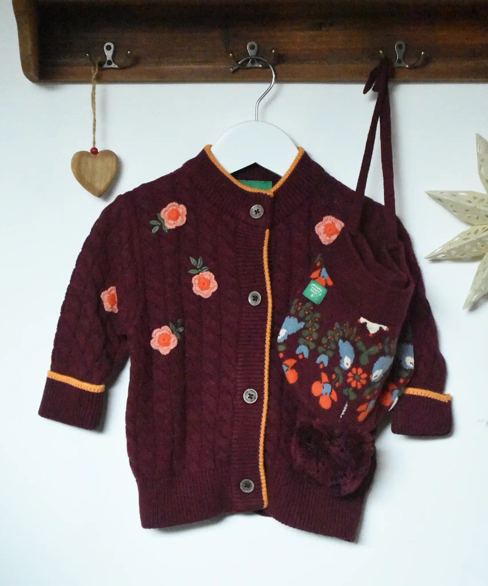Little Green Radicals organic cotton snuggly knitted cardigan with a berry petal design and yellow edges on a hanger next to a folk birds hat.