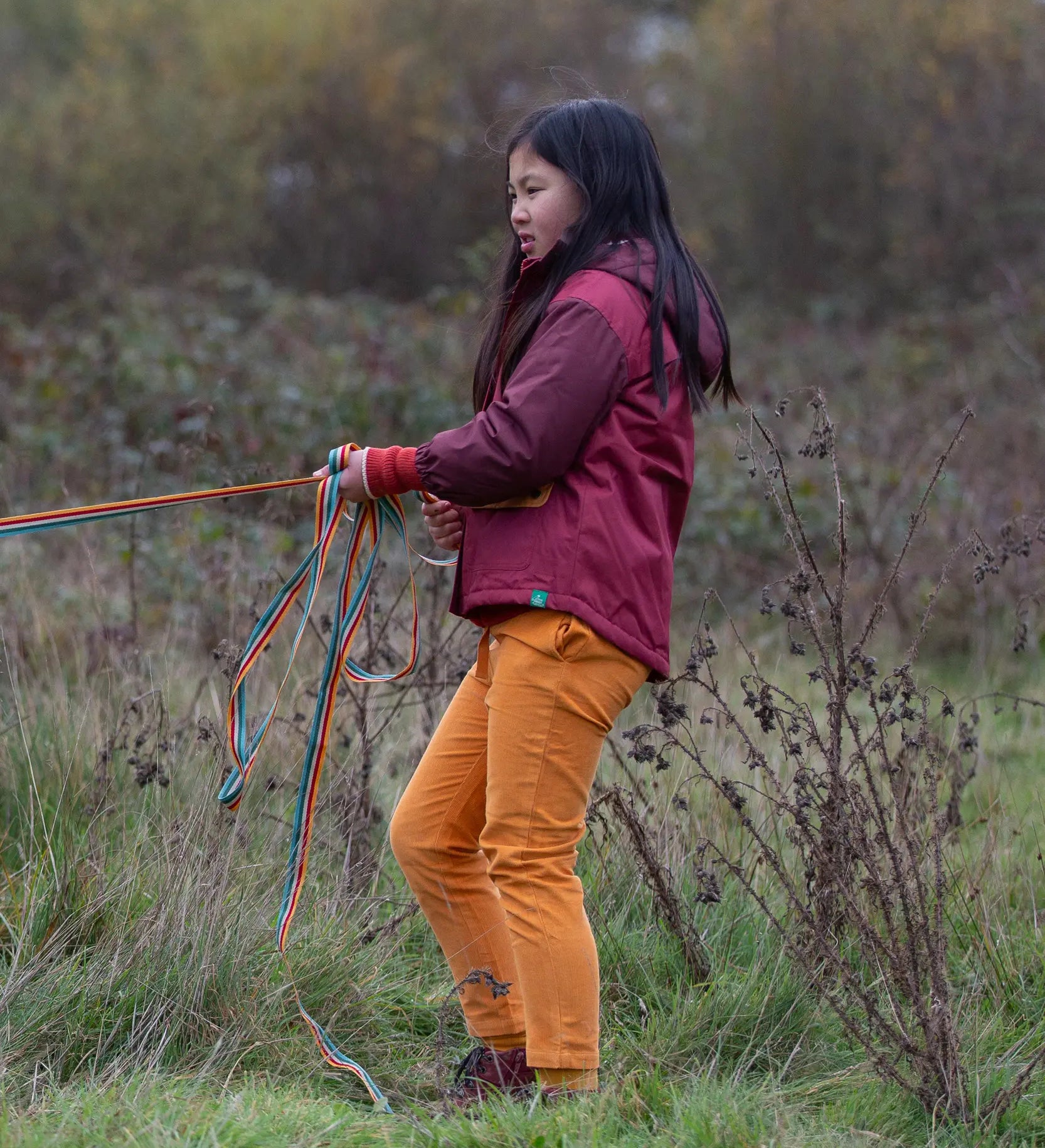 Child wearing a Little Green Radicals  berry purple recycled waterproof winter coat with yellow gold trousers.