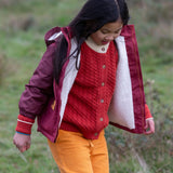 Child wearing a Little Green Radicals  berry purple recycled waterproof winter coat with a red knitted cardigan underneath and yellow gold trousers.