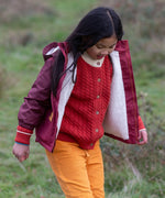 Child wearing a Little Green Radicals  berry purple recycled waterproof winter coat with a red knitted cardigan underneath and yellow gold trousers.