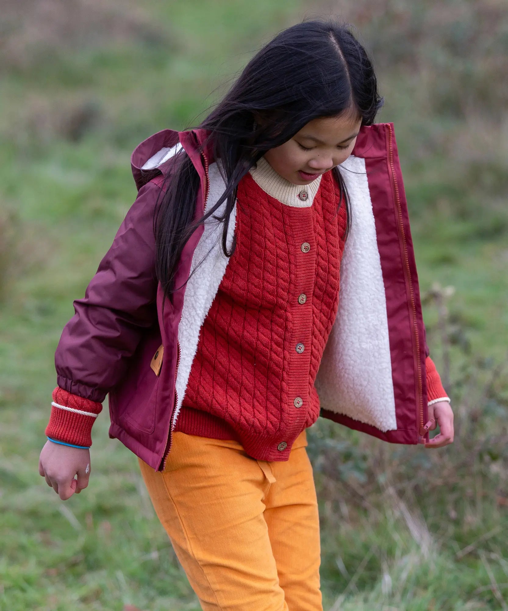 Child wearing a Little Green Radicals  berry purple recycled waterproof winter coat with a red knitted cardigan underneath and yellow gold trousers.