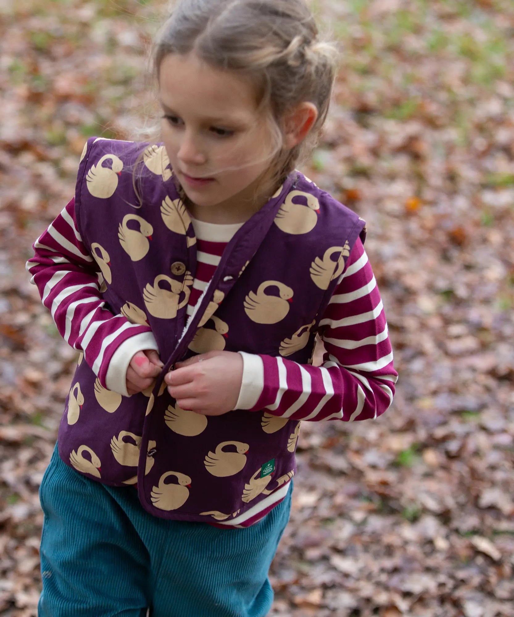 Child wearing the Little Green Radicals berry purple striped long sleeve top with green joggers and a purple sleeping swan gilet.