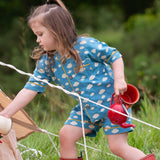 A child wearing the Little Green Radicals Blue Clouds Button Through Pyjama Short Set with red wellingtons whilst walking through long grass. The set contains a traditional style, short sleeved shirt style top with matching shorts. The print has a blue background with an over white cloud print. Little green Radicals have a wide range of organic cotton, Fairtrade children's clothing and outerwear available here at Babipur.