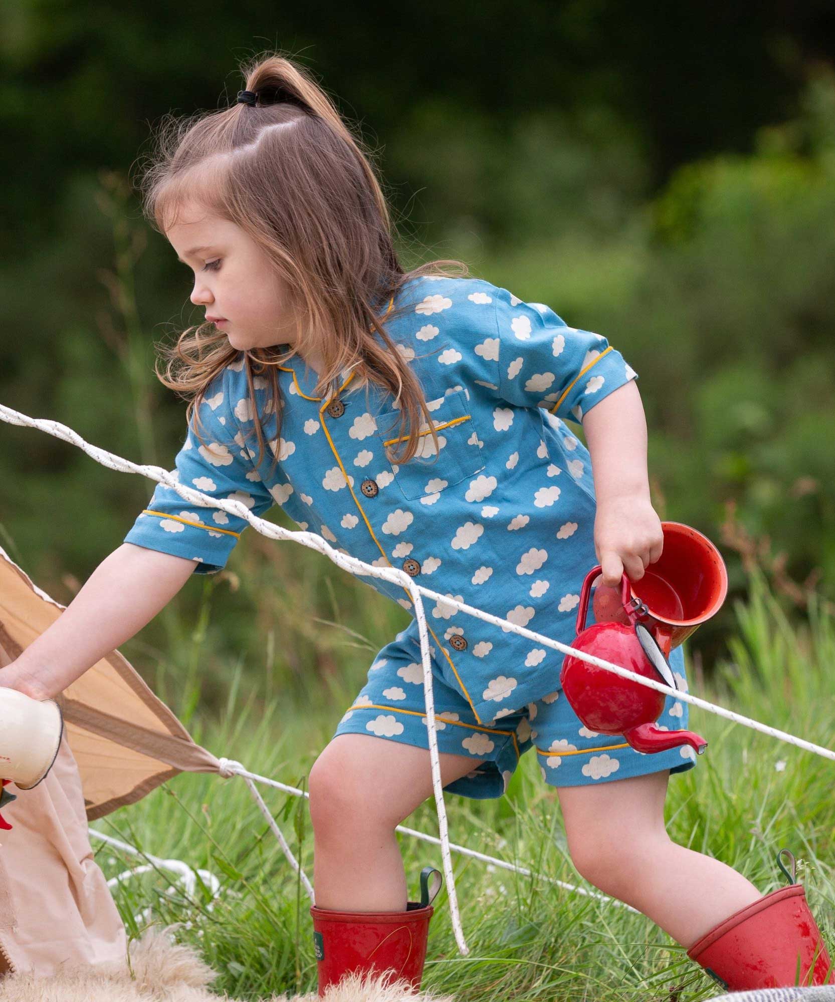 A child wearing the Little Green Radicals Blue Clouds Button Through Pyjama Short Set with red wellingtons whilst walking through long grass. The set contains a traditional style, short sleeved shirt style top with matching shorts. The print has a blue background with an over white cloud print. Little green Radicals have a wide range of organic cotton, Fairtrade children's clothing and outerwear available here at Babipur.