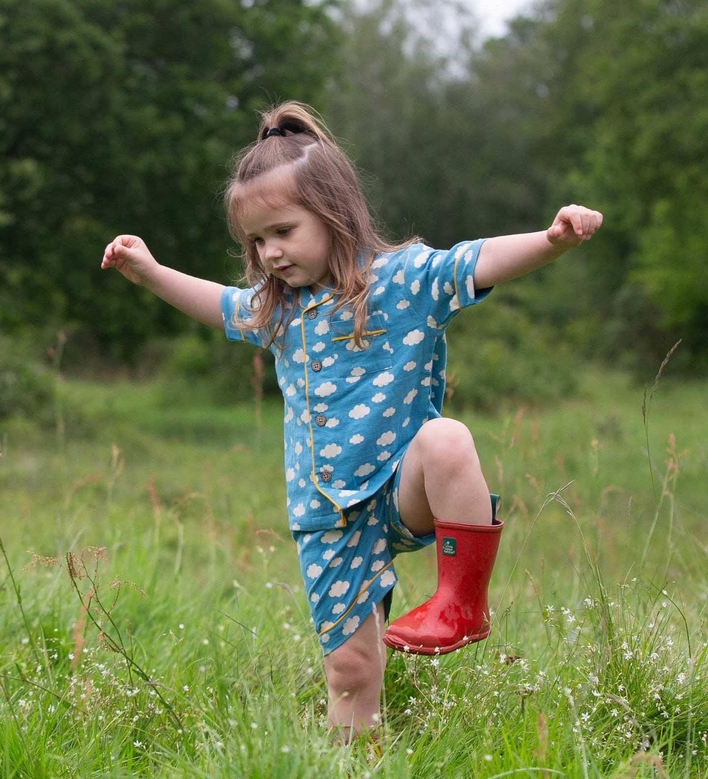 A child wearing the Little Green Radicals Blue Clouds Button Through Pyjama Short Set with red wellingtons whilst walking through long grass. The set contains a traditional style, short sleeved shirt style top with matching shorts. The print has a blue background with an over white cloud print. Little green Radicals have a wide range of organic cotton, Fairtrade children's clothing and outerwear available here at Babipur.