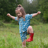 A child wearing the Little Green Radicals Blue Clouds Button Through Pyjama Short Set with red wellingtons whilst walking through long grass. The set contains a traditional style, short sleeved shirt style top with matching shorts. The print has a blue background with an over white cloud print. Little green Radicals have a wide range of organic cotton, Fairtrade children's clothing and outerwear available here at Babipur.