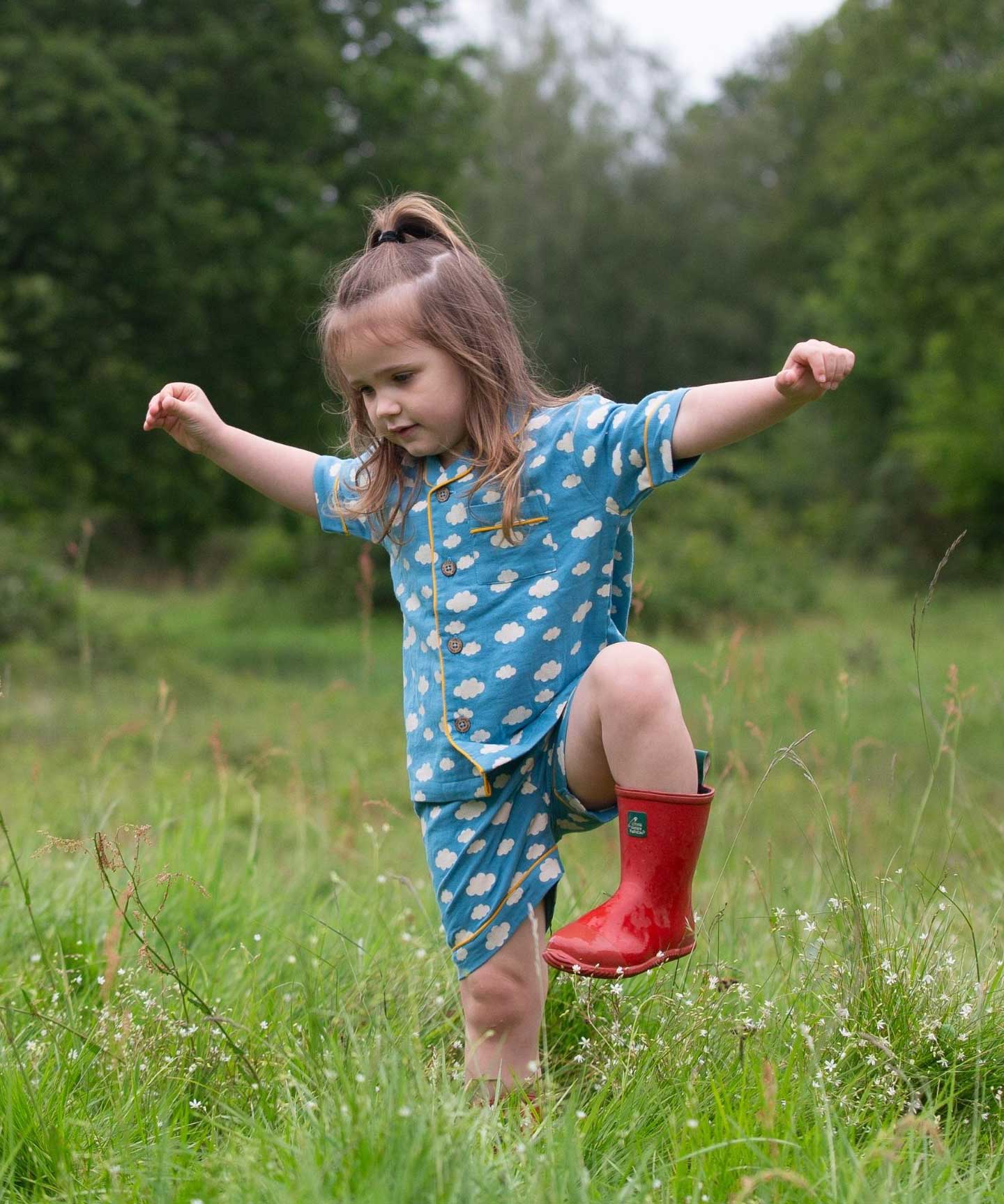 A child wearing the Little Green Radicals Blue Clouds Button Through Pyjama Short Set with red wellingtons whilst walking through long grass. The set contains a traditional style, short sleeved shirt style top with matching shorts. The print has a blue background with an over white cloud print. Little green Radicals have a wide range of organic cotton, Fairtrade children's clothing and outerwear available here at Babipur.