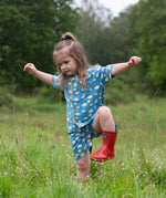 A child wearing the Little Green Radicals Blue Clouds Button Through Pyjama Short Set with red wellingtons whilst walking through long grass. The set contains a traditional style, short sleeved shirt style top with matching shorts. The print has a blue background with an over white cloud print. Little green Radicals have a wide range of organic cotton, Fairtrade children's clothing and outerwear available here at Babipur.