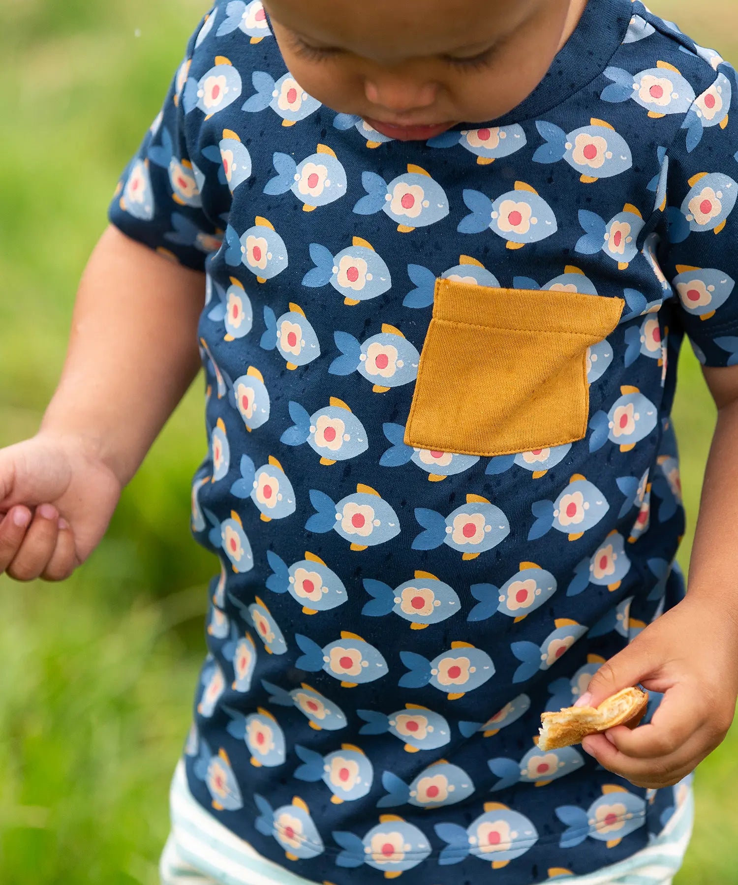 A close up of the short sleeved top from the Little Green Radicals little fish organic cotton t-shirt and jogger playset outfit. A navy top with repeat fish print and a yellow chest pocket with blue and cream striped joggers.