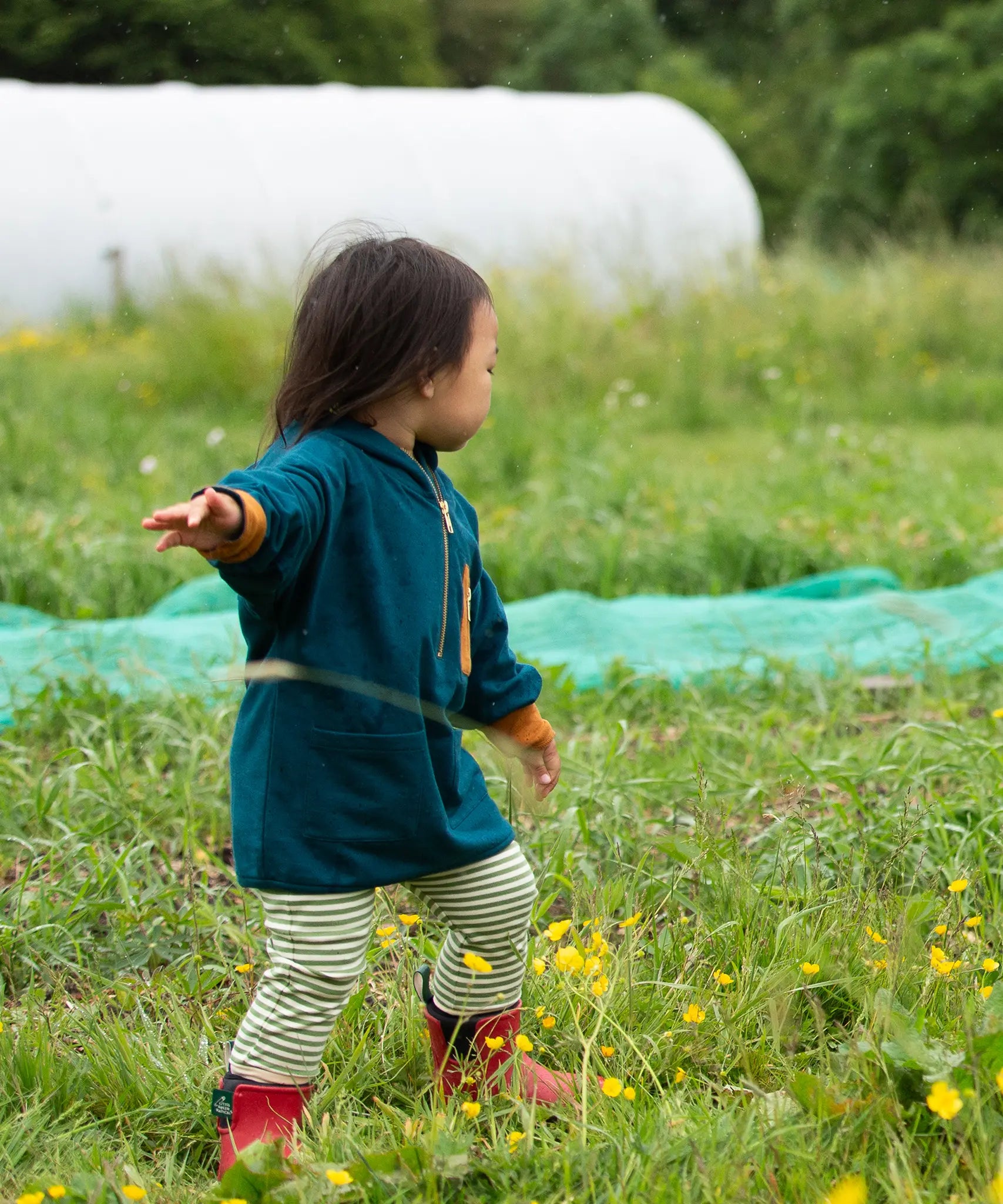 Child wearing a Little Green Radicals children's atlantic deep blue organic cotton quarter zip hoodie with striped leggings underneath and red wellington boots. 