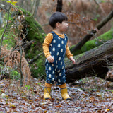 Child wearing Little Green Radicals  blue penguins everyday GOTS organic cotton dungarees, standing on a leaf covered ground in a forest. Navy blue background with an all over blue penguin print with yellow gold leg cuffs.
