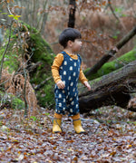 Child wearing Little Green Radicals  blue penguins everyday GOTS organic cotton dungarees, standing on a leaf covered ground in a forest. Navy blue background with an all over blue penguin print with yellow gold leg cuffs.