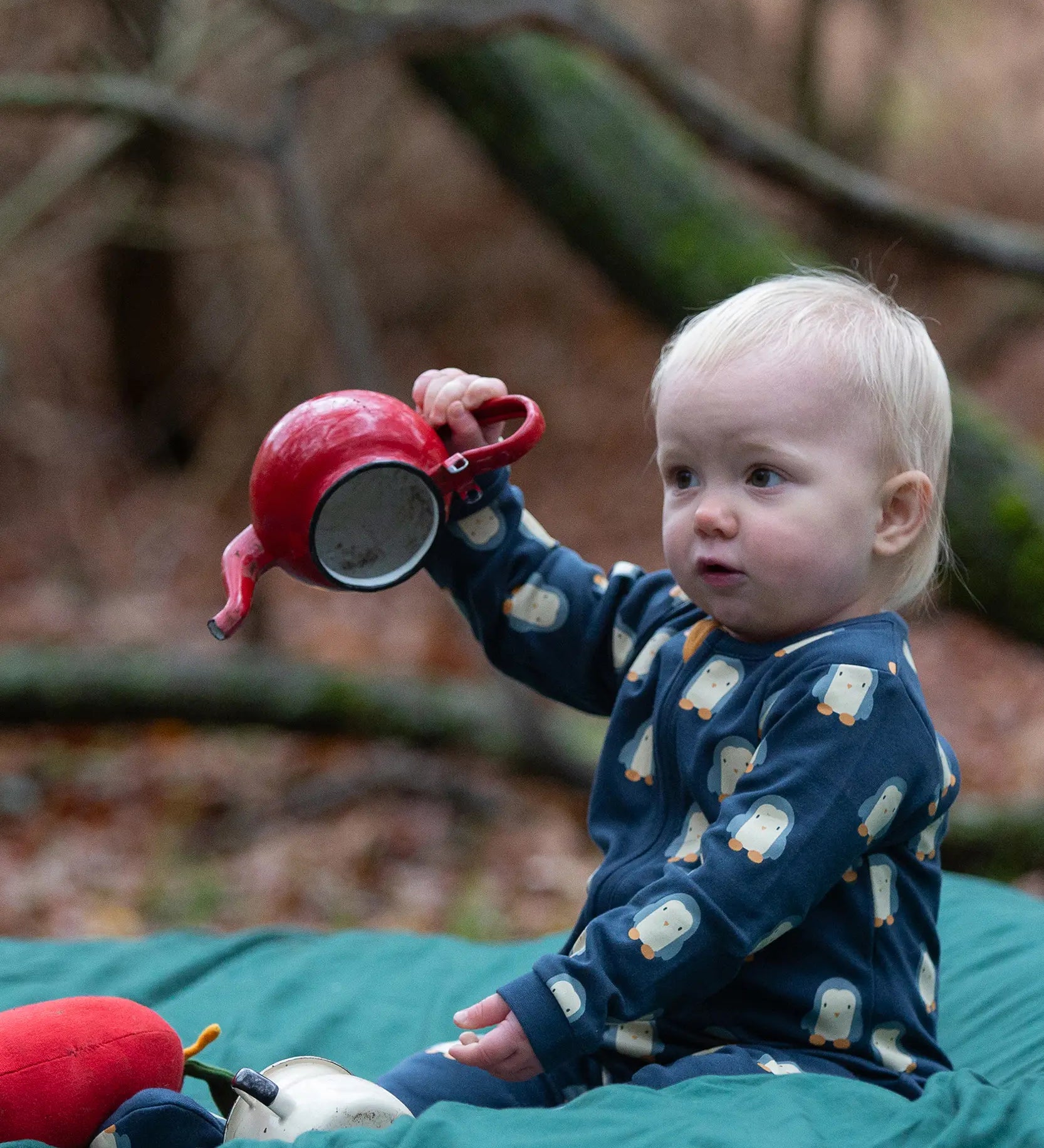 Baby wearing the Little Green Radicals blue penguins GOTS organic cotton zip up babygrow sitting on a green blanket outdoors. Navy blue coloured cotton with an all over penguin print.