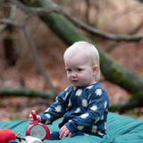 Baby wearing the Little Green Radicals blue penguins GOTS organic cotton zip up babygrow sitting on a green blanket outdoors. Navy blue coloured cotton with an all over penguin print.