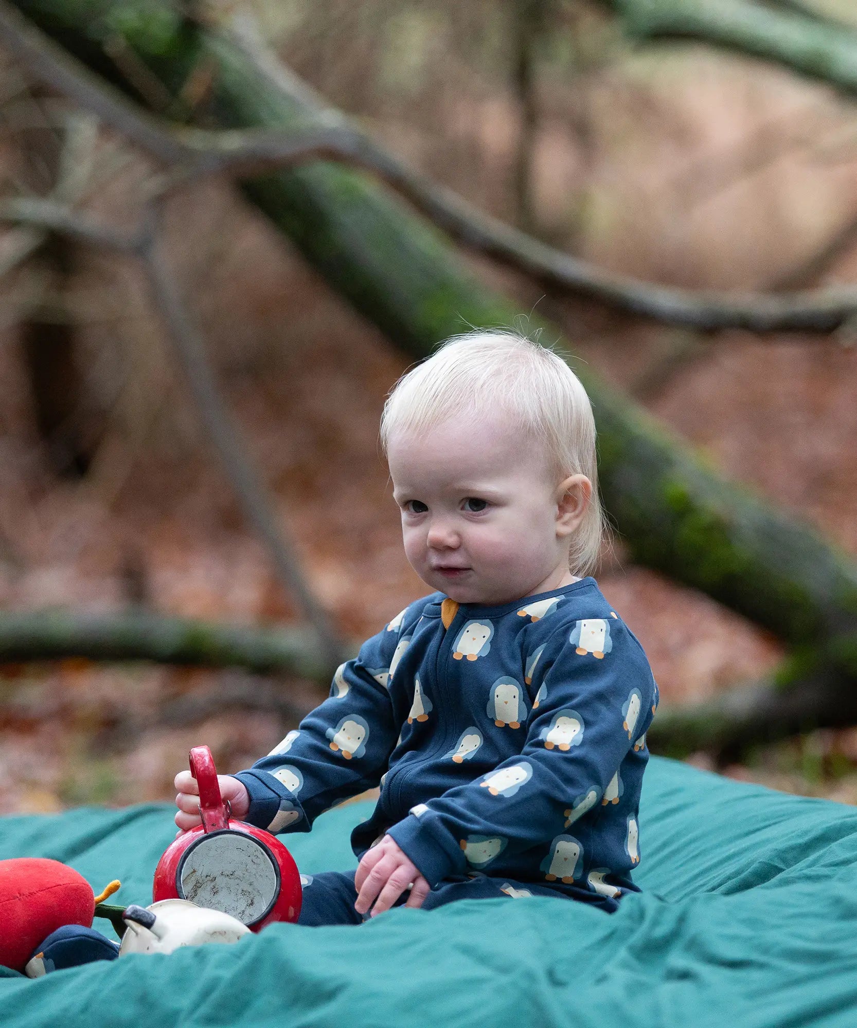 Baby wearing the Little Green Radicals blue penguins GOTS organic cotton zip up babygrow sitting on a green blanket outdoors. Navy blue coloured cotton with an all over penguin print.