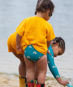 A young child wearing a Little Green Radicals Teal Rainbows UPF 50+ Recycled Reusable Swim Nappy with a yellow gold coloured t-shirt. The child is facing away from the camera.