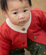 A young child wearing a Little Green Radicals From One To Another Burnt Ochre Pointelle Knitted Cardigan with green coloured shorts. Little green Radicals have a wide range of organic cotton, Fairtrade children's clothing and outerwear available here at Babipur.