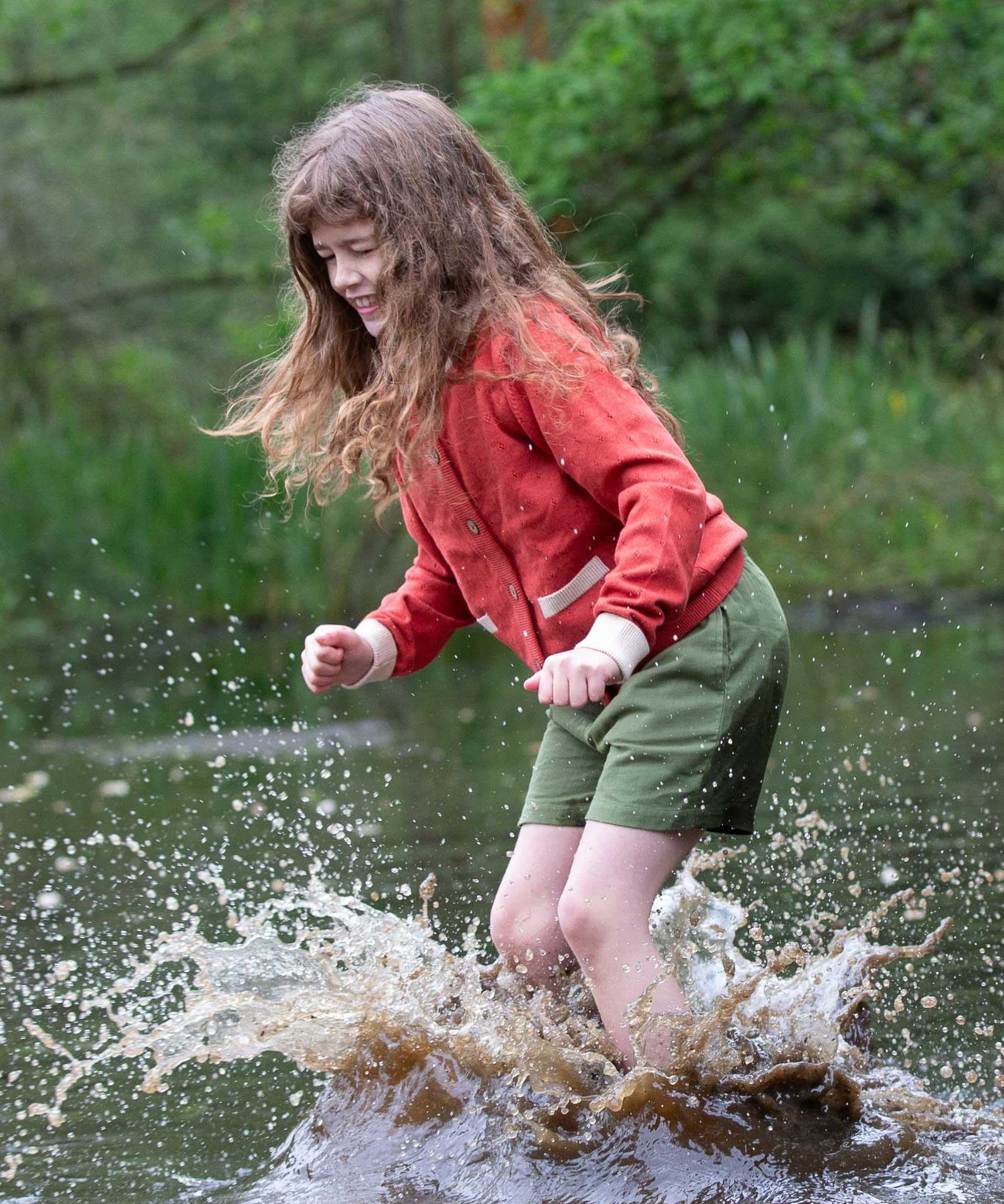 A child wearing a Little Green Radicals From One To Another Burnt Ochre Pointelle Knitted Cardigan with green coloured shorts. The child is standing in water, splashing. Little green Radicals have a wide range of organic cotton, Fairtrade children's clothing and outerwear available here at Babipur.
