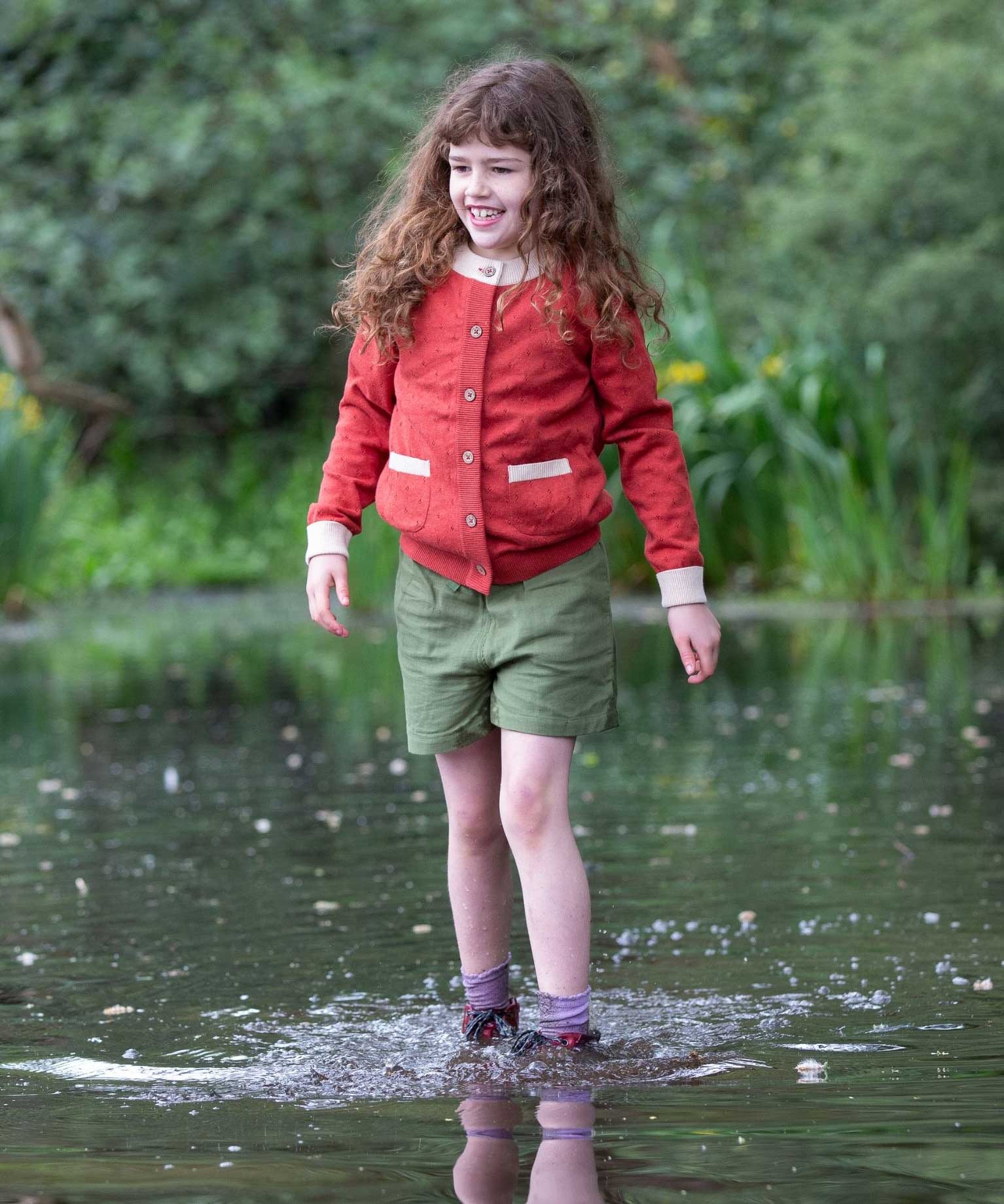 A child wearing a Little Green Radicals From One To Another Burnt Ochre Pointelle Knitted Cardigan with green coloured shorts. Little green Radicals have a wide range of organic cotton, Fairtrade children's clothing and outerwear available here at Babipur.