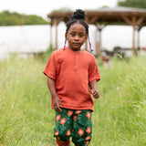 Child wearing the Little Green Radicals children's burnt ochre red short sleeved henley top with button up top and pink flower print dark green trousers.