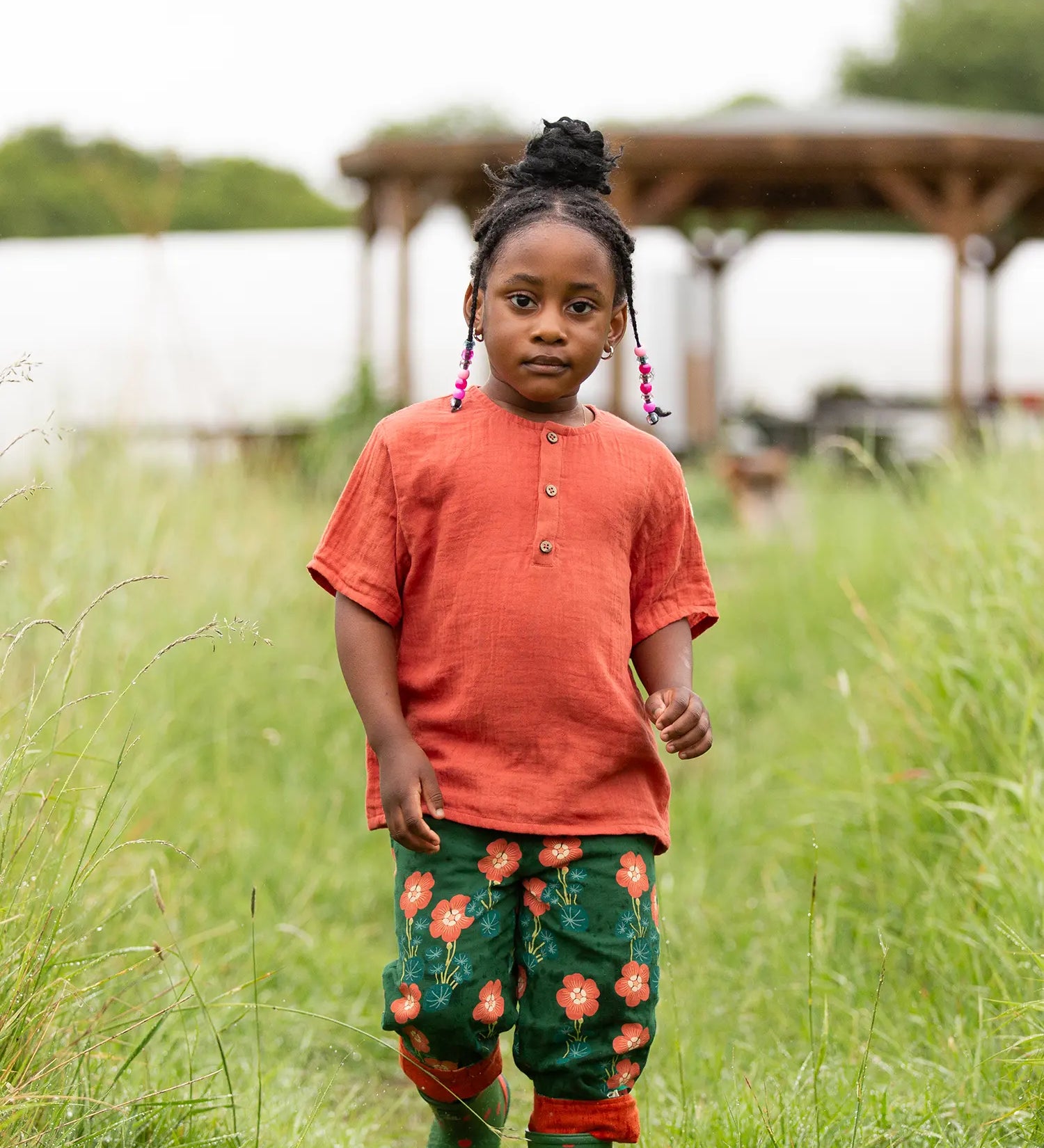Child wearing the Little Green Radicals children's burnt ochre red short sleeved henley top with button up top and pink flower print dark green trousers.