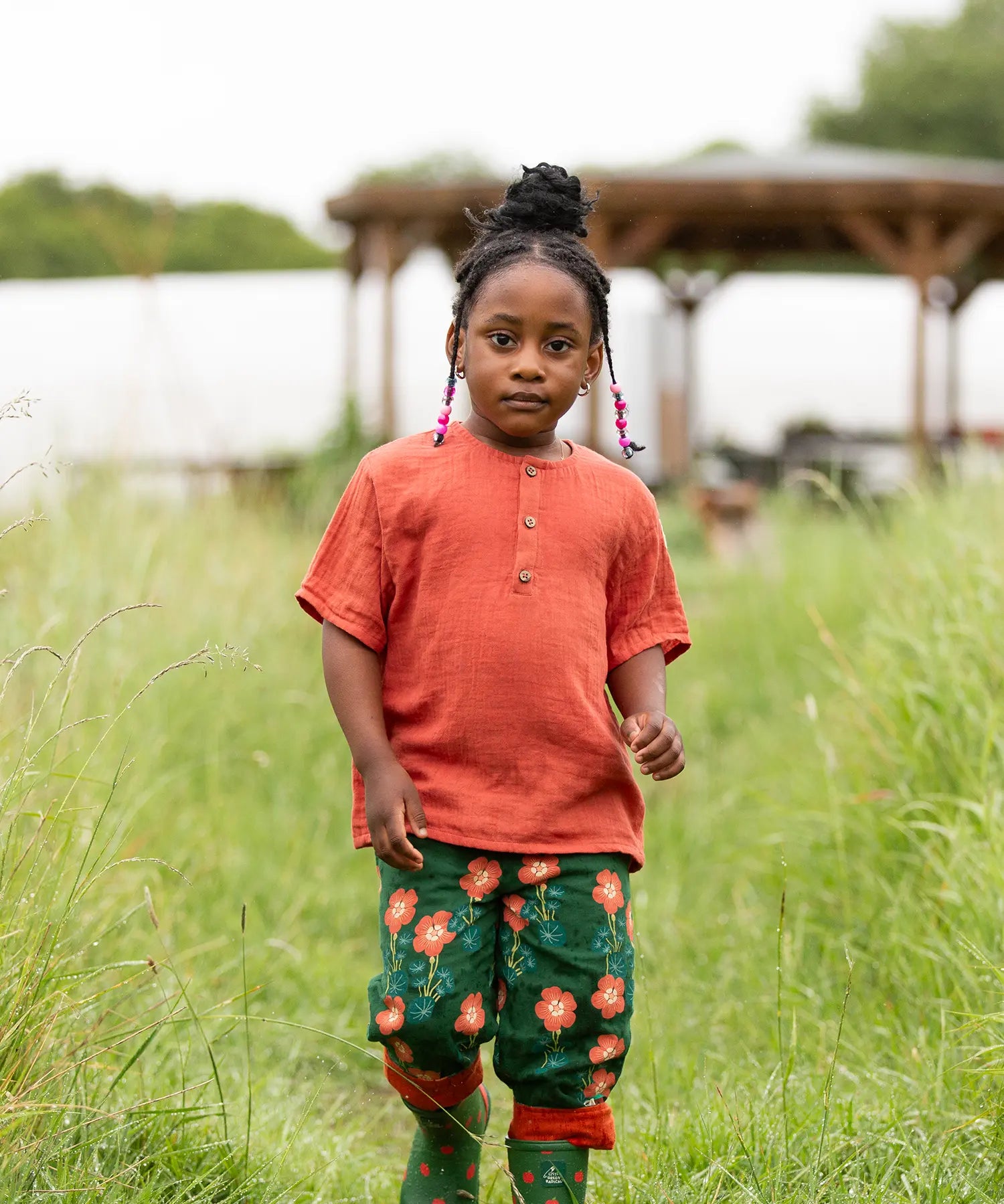 Child wearing the Little Green Radicals children's burnt ochre red short sleeved henley top with button up top and pink flower print dark green trousers.