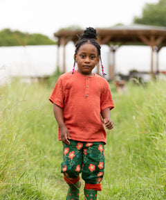 Child wearing the Little Green Radicals children's burnt ochre red short sleeved henley top with button up top and pink flower print dark green trousers.