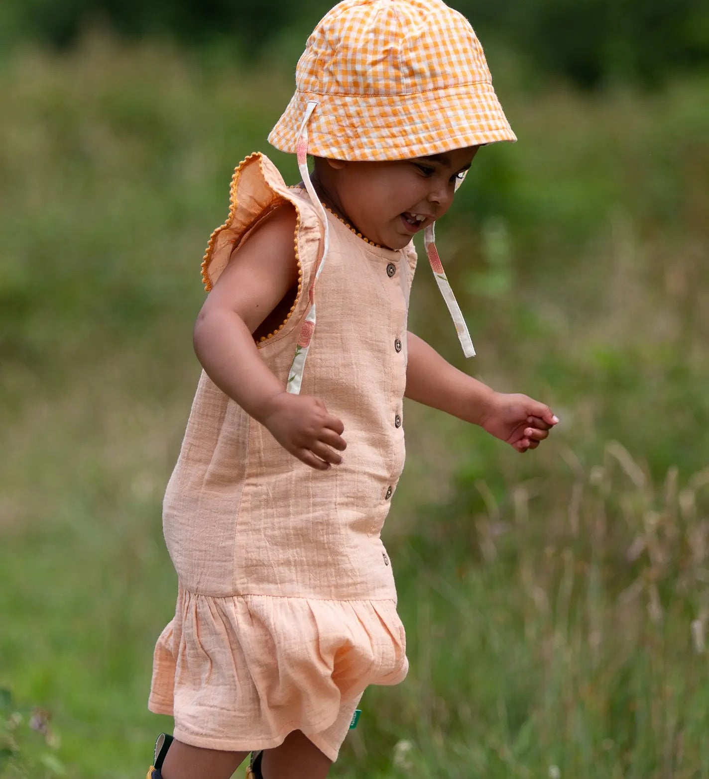 Child wearing the Little Green Radicals coral pink dropped waist children's dress with ochre yellow pom pom ribbon edging on the sleeves and collar with a yellow and white gingham sun hat.
