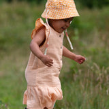 Child wearing the Little Green Radicals coral pink dropped waist children's dress with ochre yellow pom pom ribbon edging on the sleeves and collar with a yellow and white gingham sun hat.