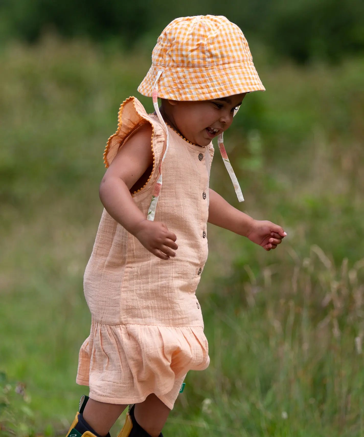 Child wearing the Little Green Radicals coral pink dropped waist children's dress with ochre yellow pom pom ribbon edging on the sleeves and collar with a yellow and white gingham sun hat.