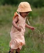 Child wearing the Little Green Radicals coral pink dropped waist children's dress with ochre yellow pom pom ribbon edging on the sleeves and collar with a yellow and white gingham sun hat.