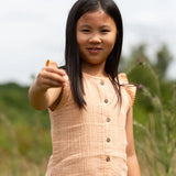 Close up of a child wearing the Little Green Radicals coral pink dropped waist button up front children's dress with ochre yellow pom pom ribbon edging on the sleeves and collar.