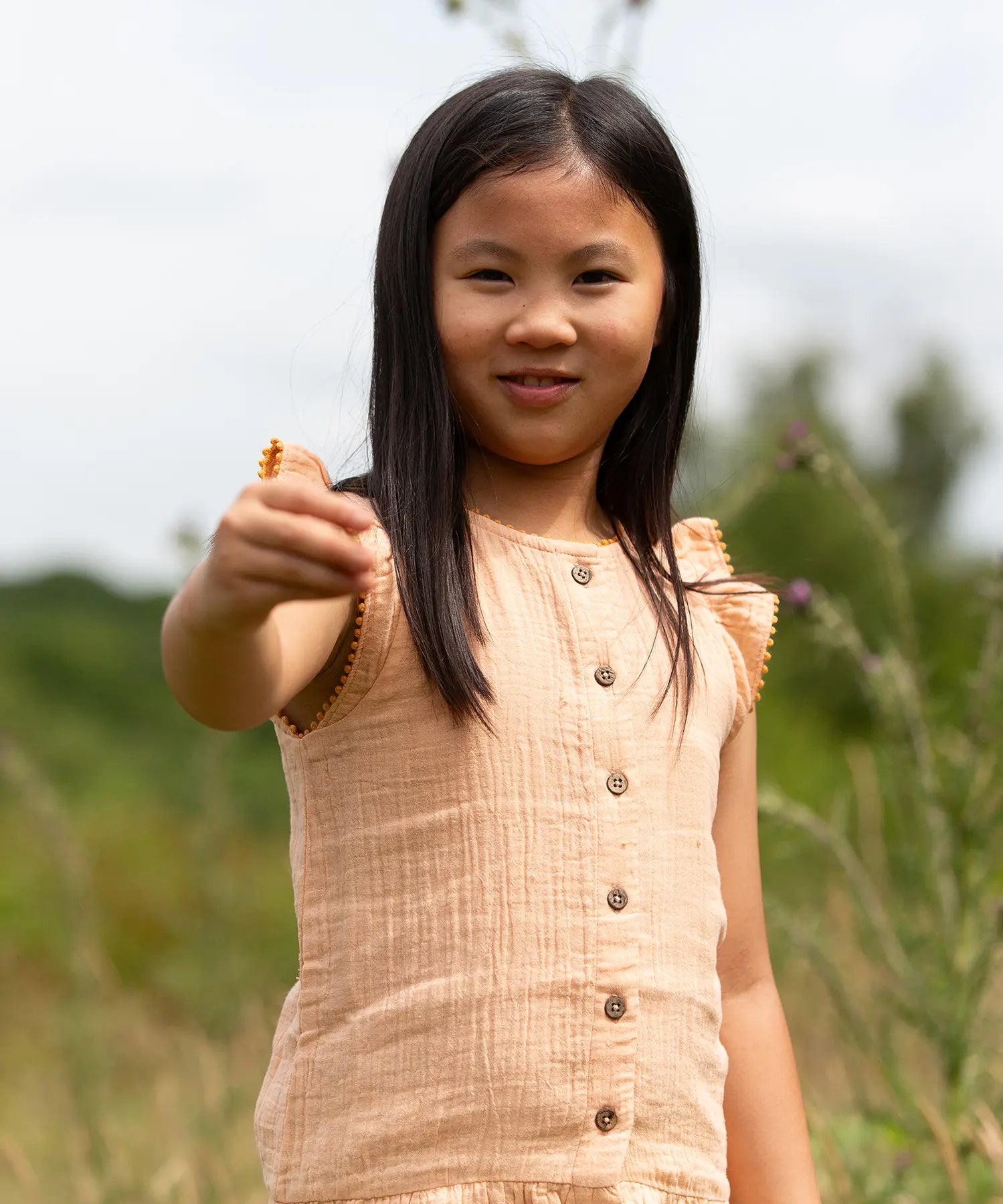 Close up of a child wearing the Little Green Radicals coral pink dropped waist button up front children's dress with ochre yellow pom pom ribbon edging on the sleeves and collar.
