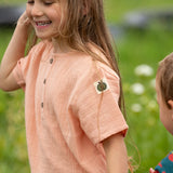 Close up of the apple tag on the arm of the Little Green Radicals kids light coral pink short sleeved henley top being worn by a child.