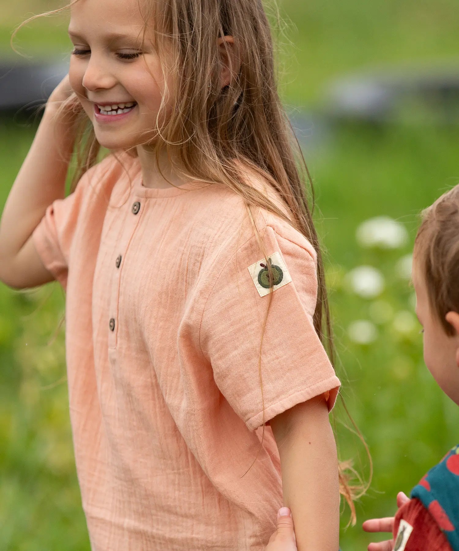 Close up of the apple tag on the arm of the Little Green Radicals kids light coral pink short sleeved henley top being worn by a child.