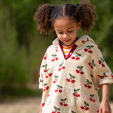 Child wearing the Little Green Radicals little cherries cream organic cotton kids hooded towel poncho.The poncho has a popper closure on the neck and a kangaroo pocket on the front. 