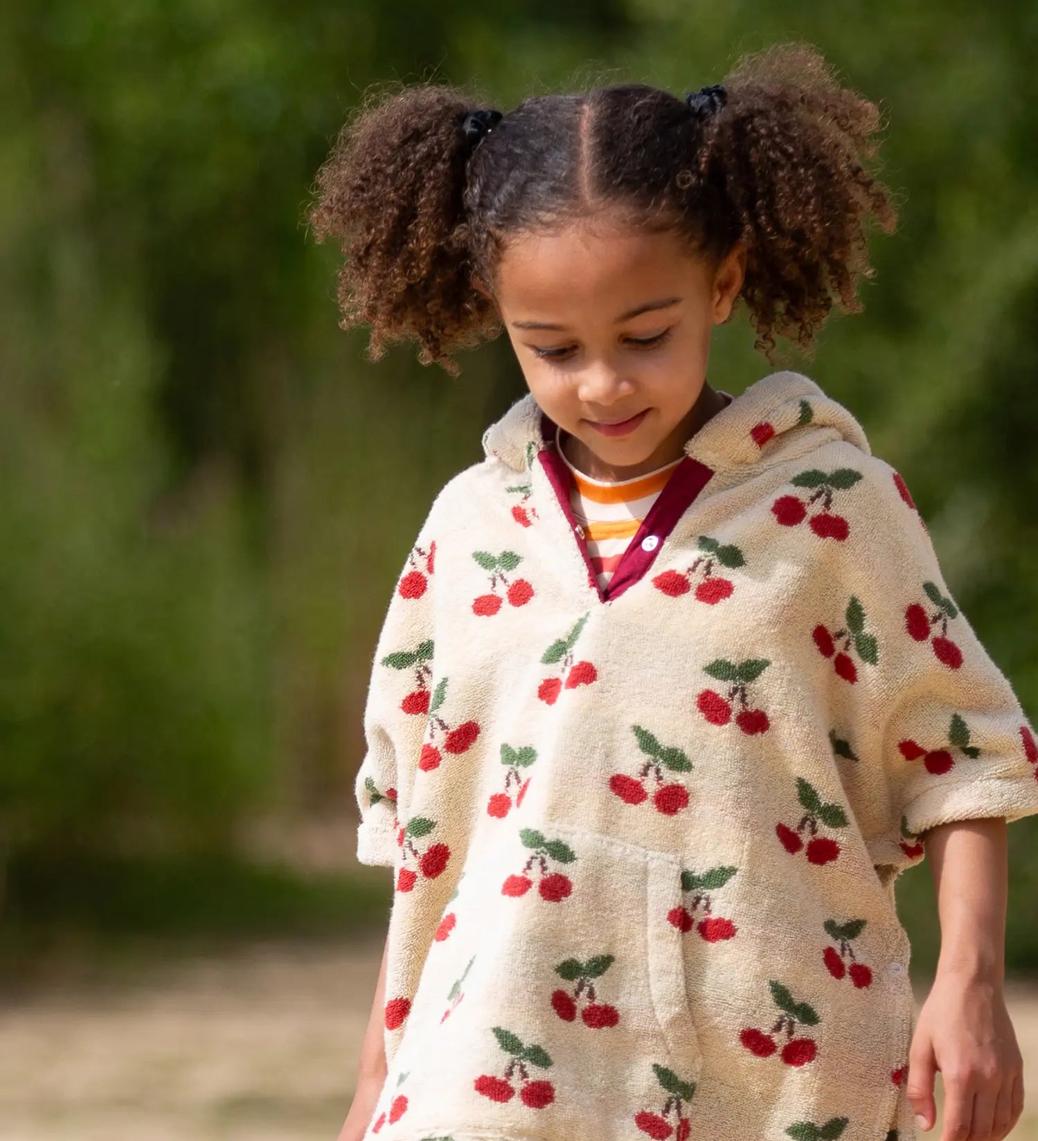 Child wearing the Little Green Radicals little cherries cream organic cotton kids hooded towel poncho.The poncho has a popper closure on the neck and a kangaroo pocket on the front. 