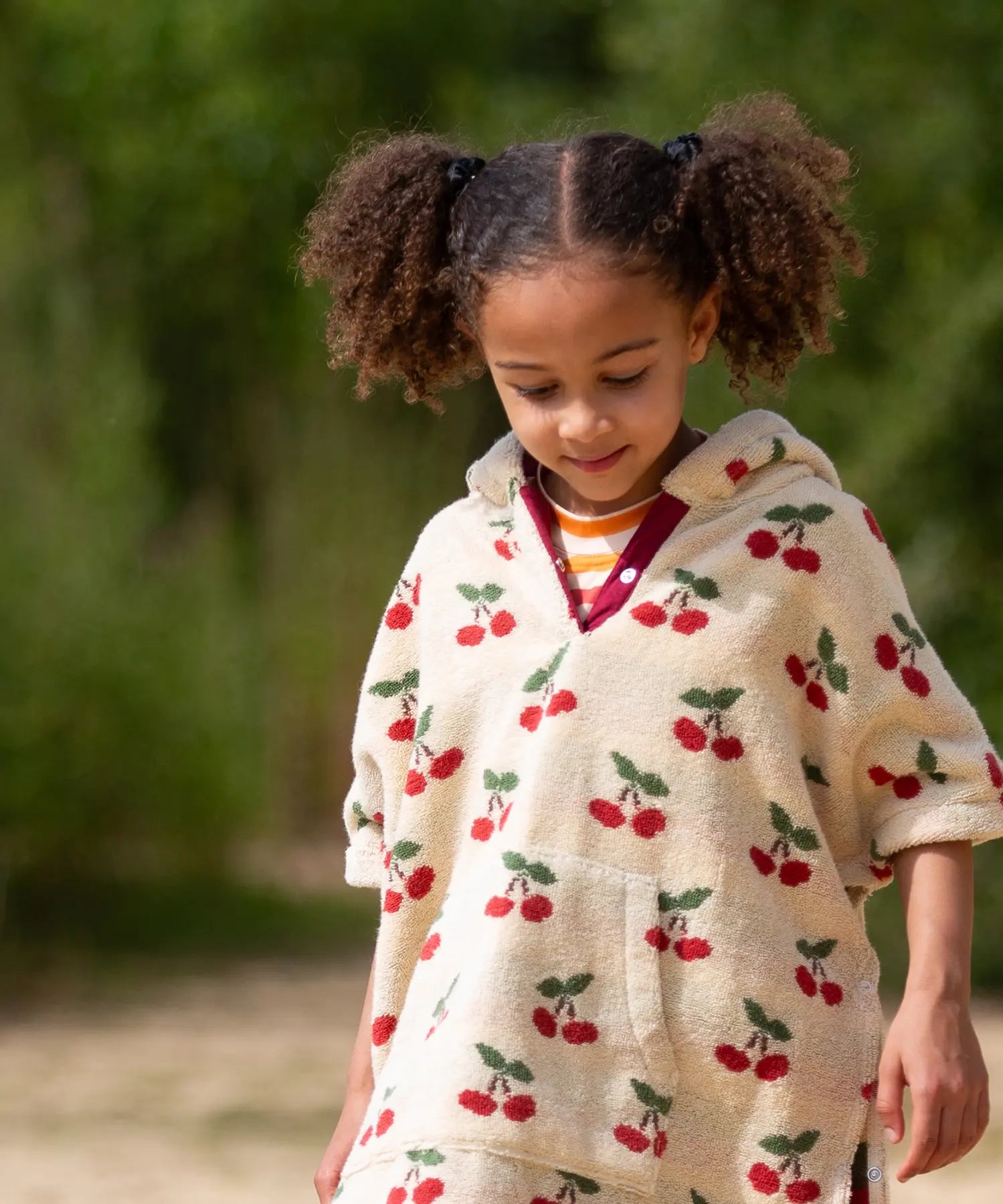 Child wearing the Little Green Radicals little cherries cream organic cotton kids hooded towel poncho.The poncho has a popper closure on the neck and a kangaroo pocket on the front. 