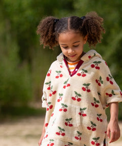 Child wearing the Little Green Radicals little cherries cream organic cotton kids hooded towel poncho.The poncho has a popper closure on the neck and a kangaroo pocket on the front. 