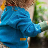 A view of the side of a child wearing the Little Green Radicals From One To Another Deep Blue Colour Block Knitted Cardigan. This organic cotton cardigan is part of a wide range of organic cotton, Fairtrade children's clothing and outerwear available here at Babipur.
