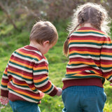 Two children wearing the Little Green Radicals knitted snuggly jumper in a  hazel rainbow striped design  with blue corduroy trousers. 